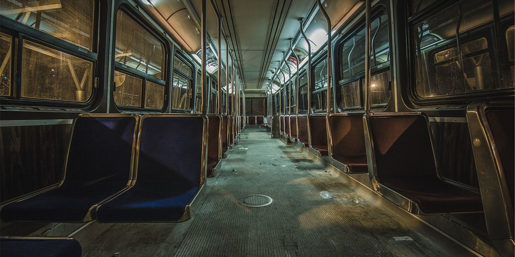 An empty, dimly lit city bus with stained floors, reflecting overhead lights and scattered trash, shot from a low central perspective.