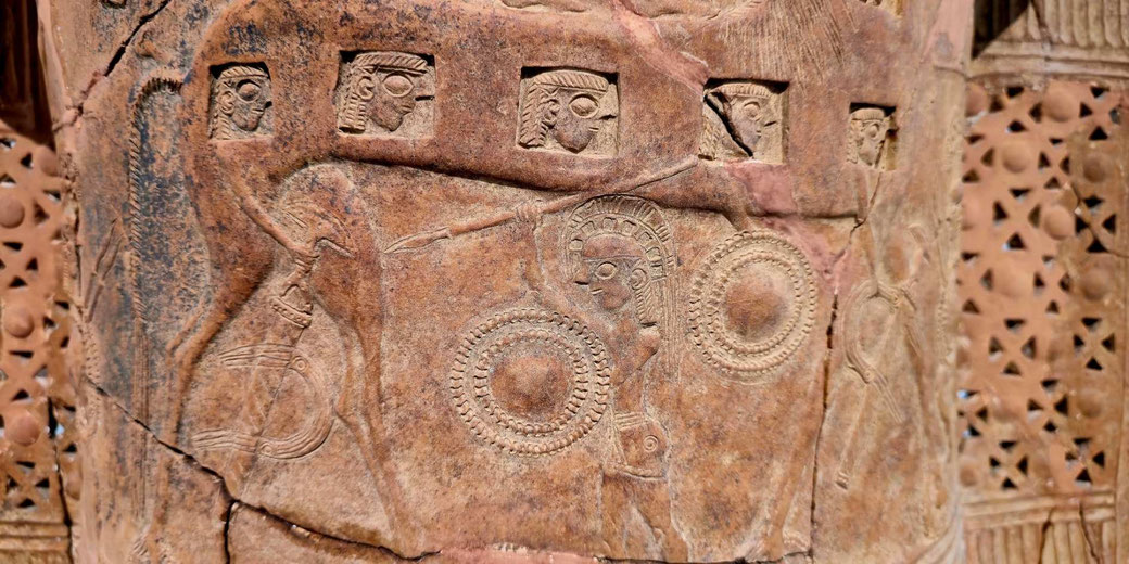 Ancient terracotta relief on the Mykonos Vase with carved warriors carrying round shields and spears beneath a row of stylised human faces.