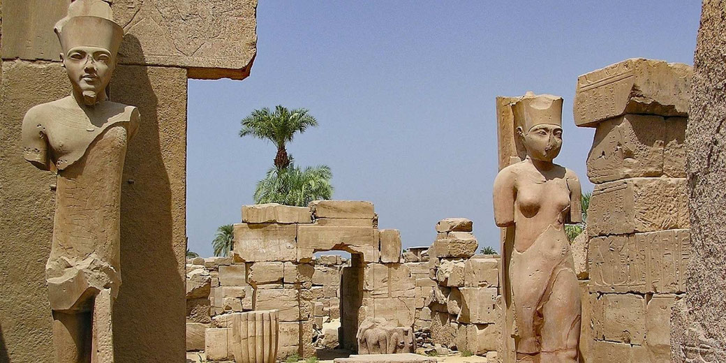 Ancient Egyptian temple site with damaged pharaoh statues, carved blocks, palm trees, and clear sky in a desert archaeological setting.
