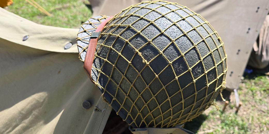 A WWII-era American M1 helmet with a net cover and a leather chinstrap, resting on military gear near a canvas tent, likely part of a historical reenactment or museum display.