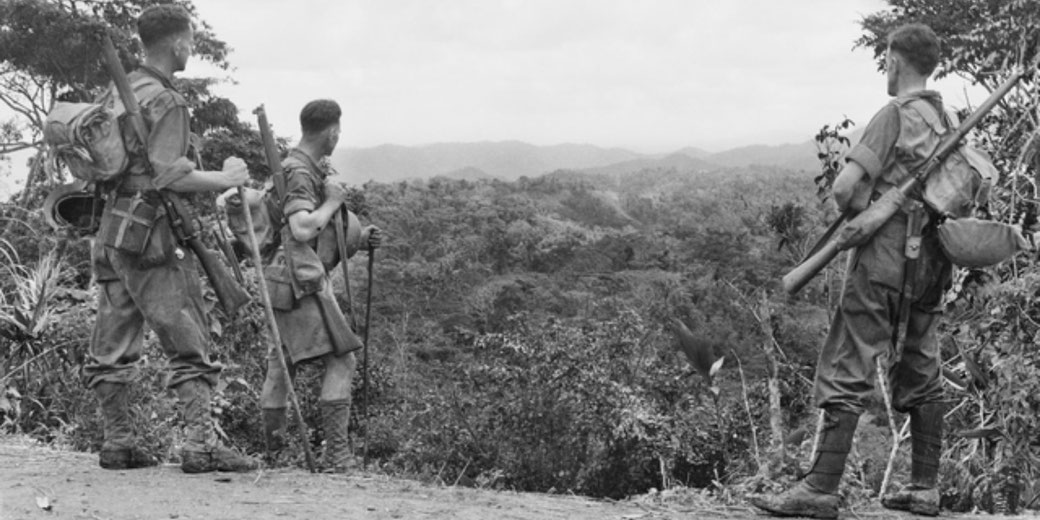 Australian soldiers pause on a ridge along the Kokoda Track, observing mountainous terrain. The image captures the difficult landscape faced during the 1942 campaign.