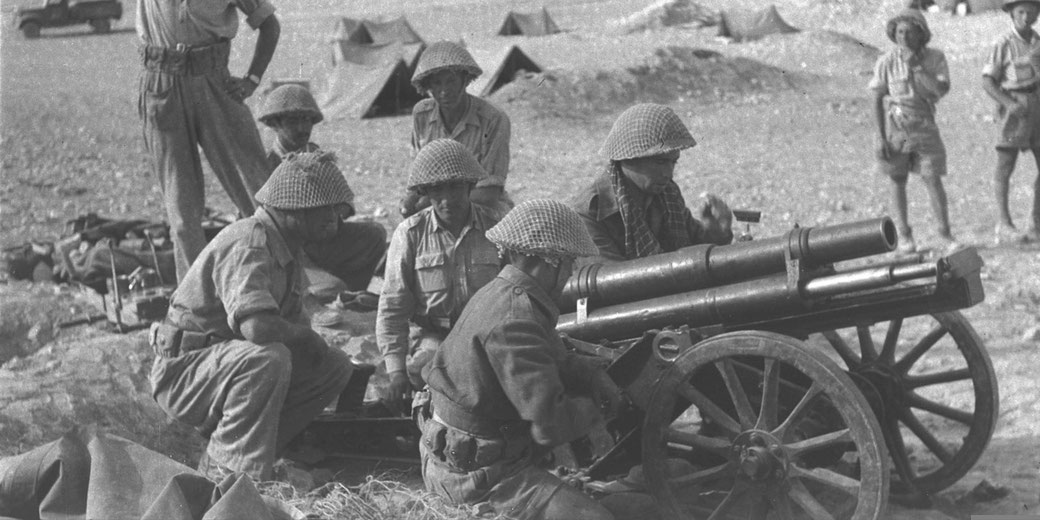 Soldiers operate a field artillery gun in a desert camp surrounded by tents.