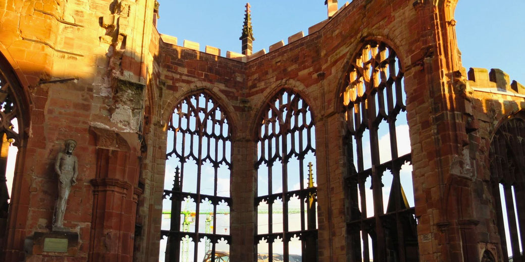 Sunlight hits the weathered stone and large gothic windows of a partially ruined cathedral wall with intricate tracery and arched frames.