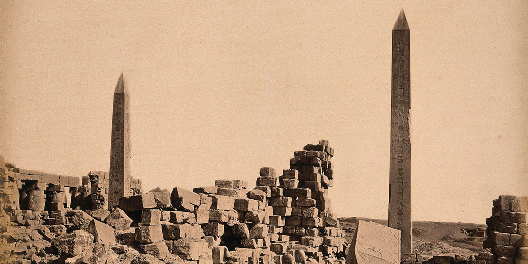 Two ancient Egyptian obelisks rise among weathered stone ruins under a clear sky in a sepia-toned image.