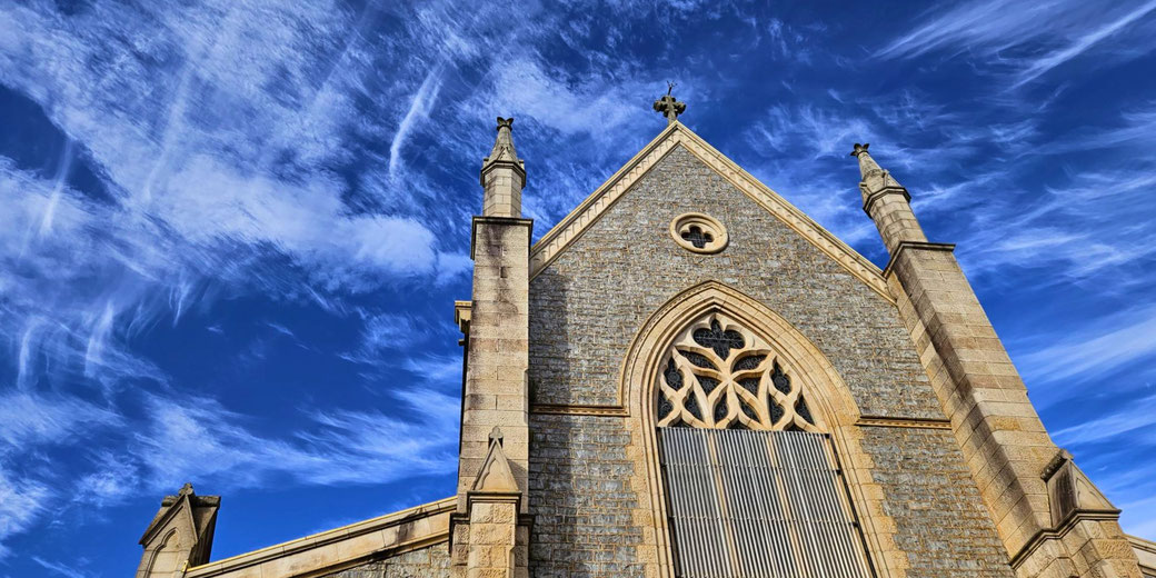A historic stone church with Gothic architectural details, pointed arches, and ornate tracery, set against a vibrant blue sky with wispy clouds.