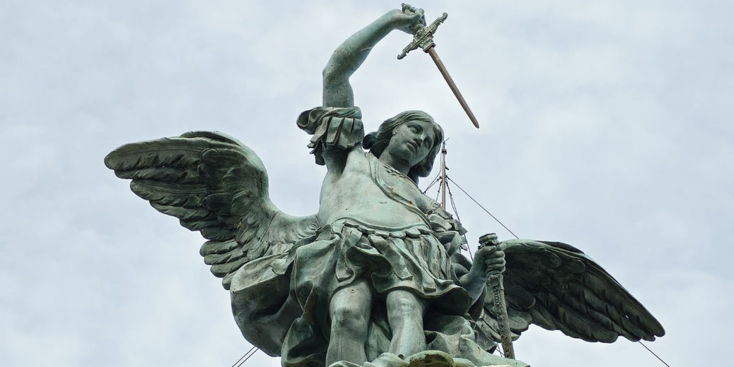 Bronze statue of a winged angel, likely Archangel Michael, raising a sword triumphantly with a calm expression against a cloudy sky.