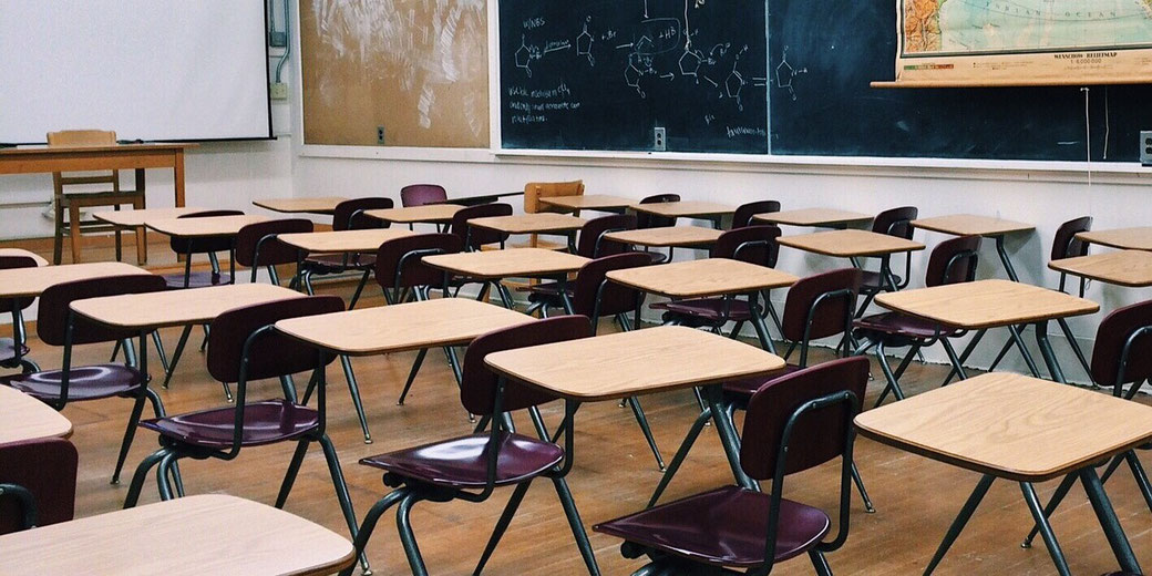 An empty classroom with rows of wooden desks, maroon chairs, a blackboard with chemistry notes, and a pull-down map.