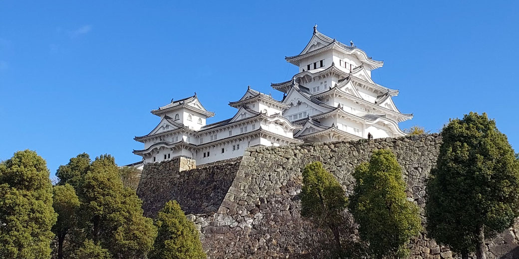 Himeji Castle, a historic Japanese castle with white walls and elegant multi-tiered roofs, sitting atop a stone foundation surrounded by lush greenery under a clear blue sky.