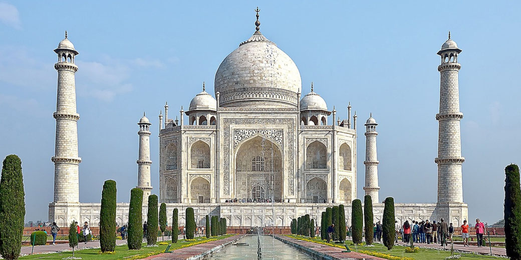 The Taj Mahal stands in bright daylight with its white marble dome, four minarets, and reflecting pool framed by green gardens.