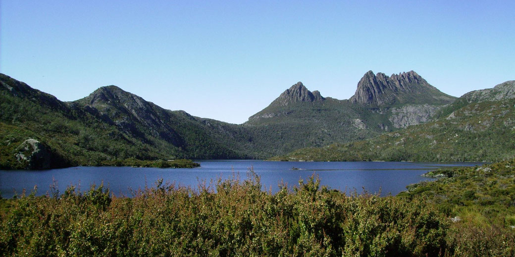 A stunning view of Cradle Mountain in Tasmania, with its jagged peaks rising above a serene blue lake, surrounded by lush greenery and under a clear, bright sky.