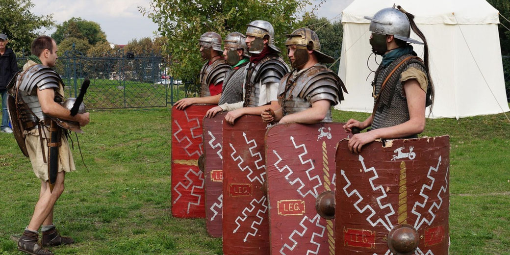 A reenactment of Roman soldiers in training. They wear segmented armor, hold rectangular shields with "LEG" markings, and stand in formation while an officer instructs them outdoors.