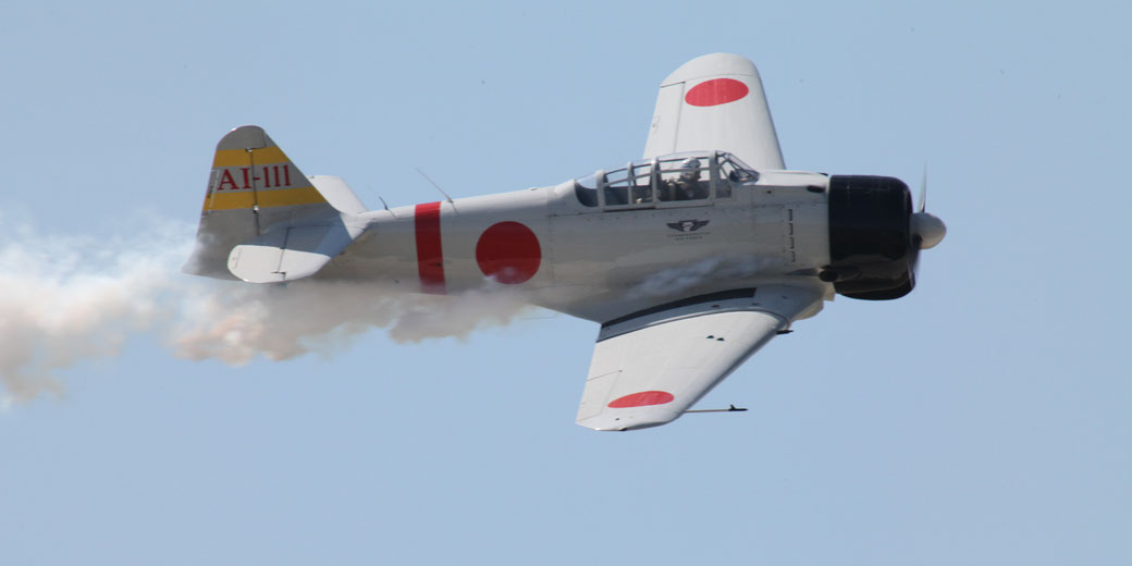 A vintage Japanese WWII aircraft with red circle insignias flies against a clear blue sky, trailing smoke.