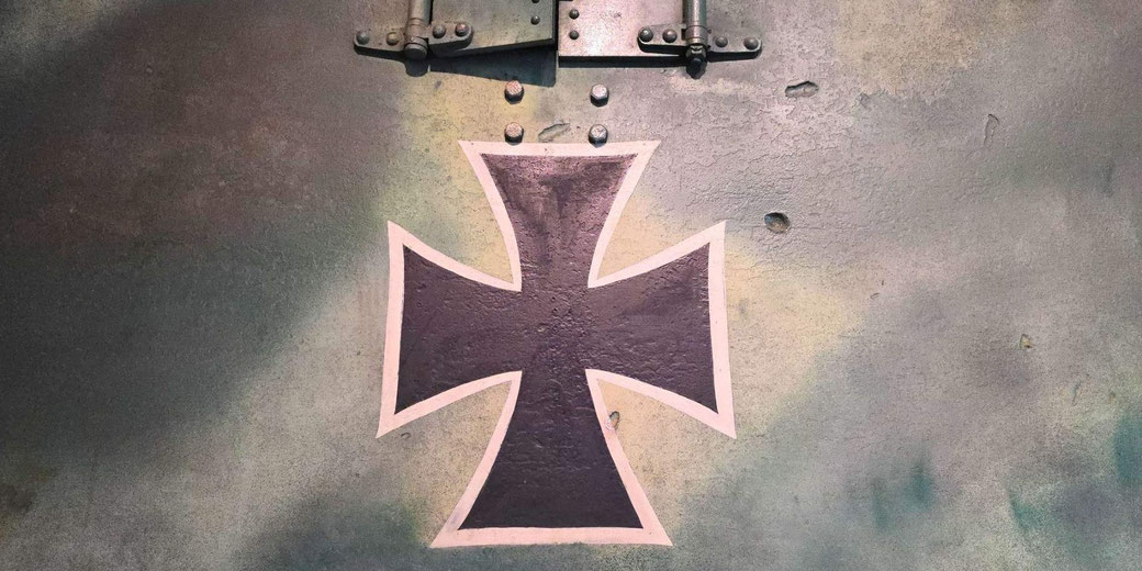 A black Iron Cross symbol with a white outline is painted on a weathered metal surface with bolts and hinges visible at the top.