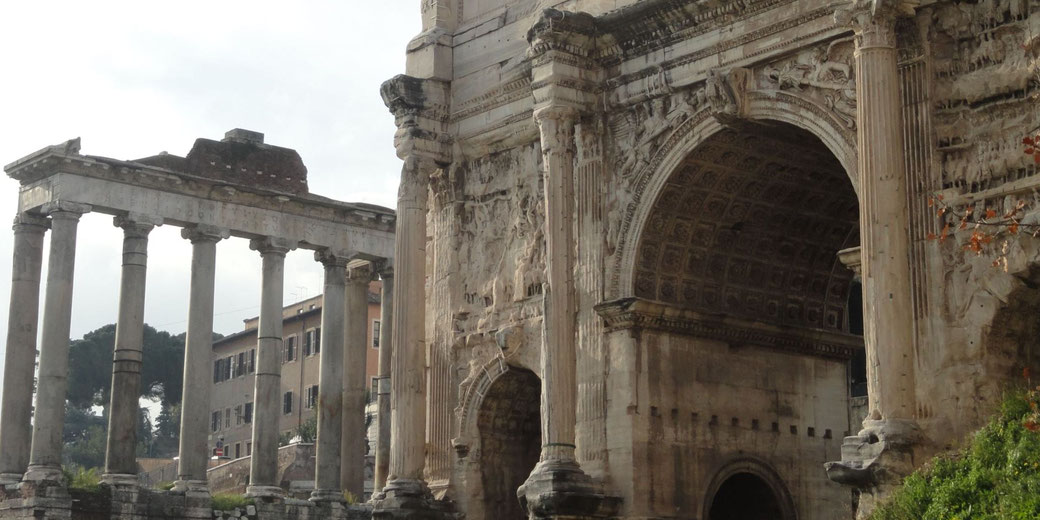 Ancient Roman ruins featuring a large triumphal arch and surviving temple columns, showcasing detailed carvings and classical architecture.