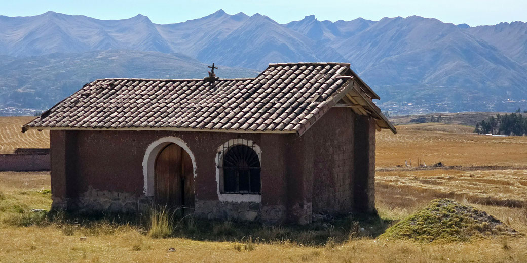 A small adobe chapel with a tiled roof stands alone in a dry field, backed by rugged mountain peaks under a clear sky.