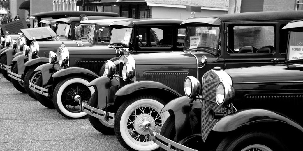 A row of polished vintage cars is lined up at a car show, captured in black and white to highlight their classic design and chrome details.