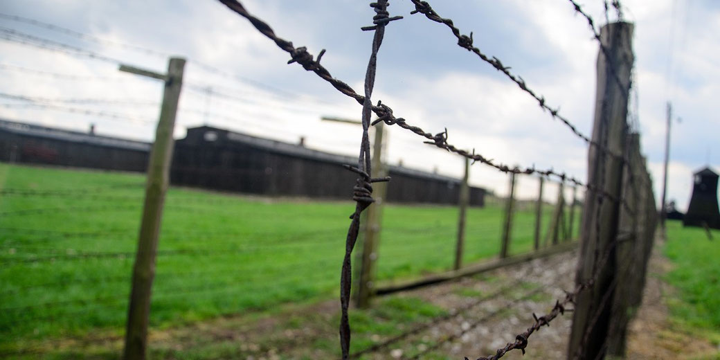 Close-up of barbed wire fence with grassy field and dark wooden buildings in the background under a cloudy sky.