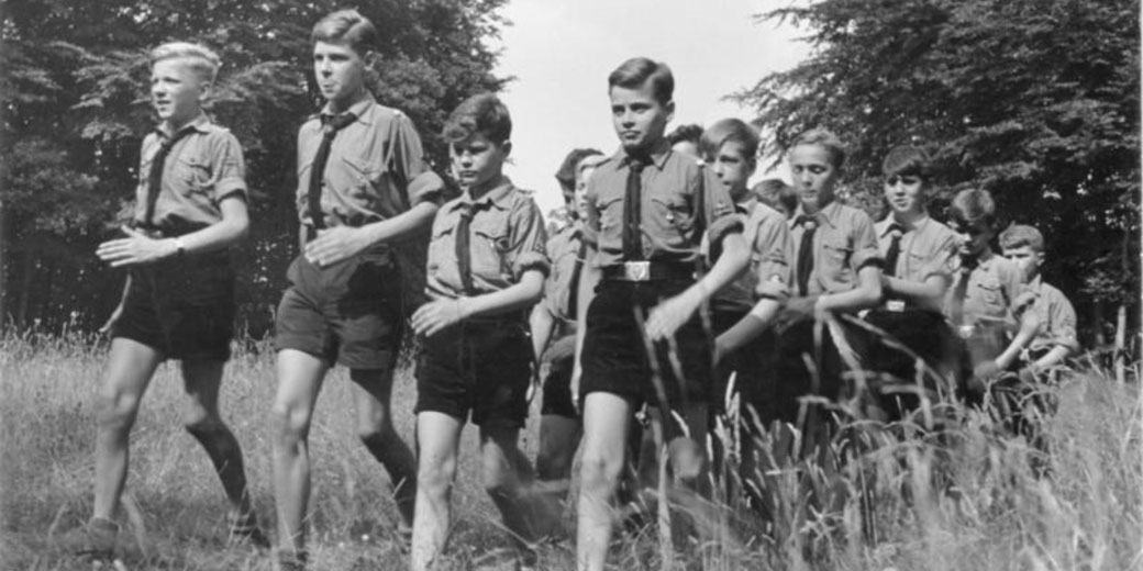 A group of boys in uniform march in formation through a grassy field, with trees in the background, captured in black and white.