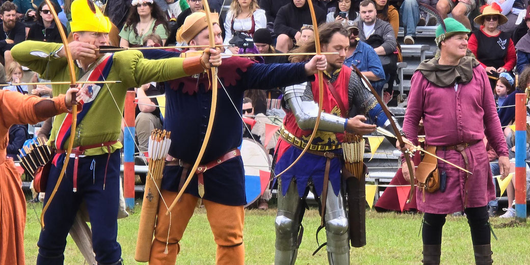 A group of archers dressed in medieval attire, each holding a bow and aiming at targets. The scene is set at a festival or reenactment, with a crowd in the background and colorful flags around the field.