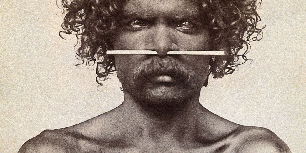 A black-and-white portrait of an Indigenous Australian man with curly hair and a mustache. He has a piercing through his nose with a long, straight object inserted horizontally.