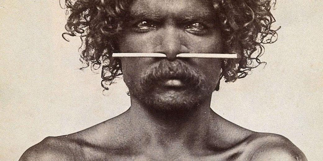 A black-and-white portrait of an Indigenous Australian man with curly hair and a mustache. He has a piercing through his nose with a long, straight object inserted horizontally.