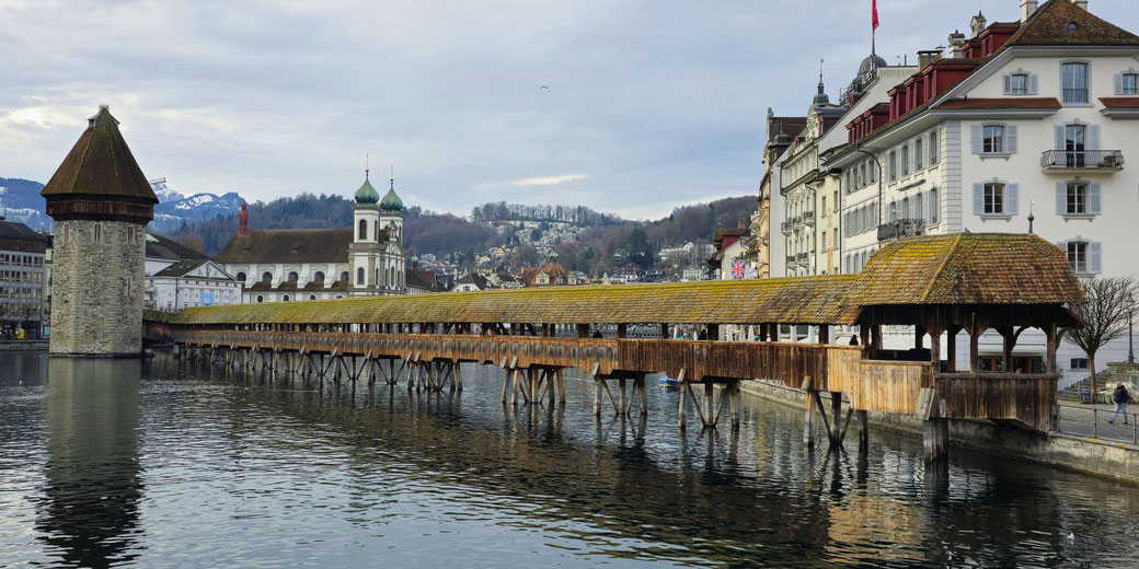The iconic wooden Chapel Bridge and Water Tower in Lucerne, Switzerland, set against a backdrop of historic buildings, a church, and snow-capped mountains under a cloudy sky.