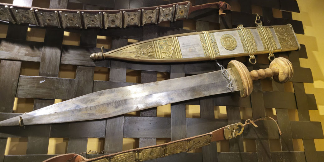 A Roman gladius with a broad, double-edged blade and a wooden hilt is shown beside its decorated scabbard, featuring engraved metalwork and suspension rings.