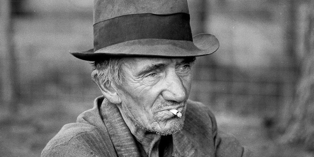 An elderly man with a weathered face smokes a cigarette, wearing a tilted hat and coat, captured in black and white.