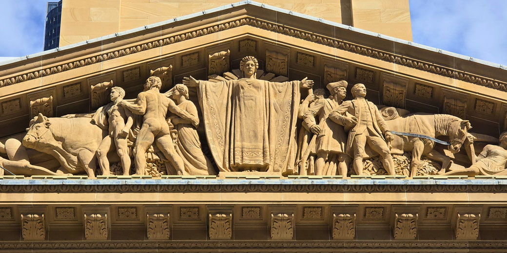 An ornate stone relief on a building facade features a central robed figure with outstretched arms, flanked by farmers, animals, and settlers, symbolizing prosperity, labor, and civilization.
