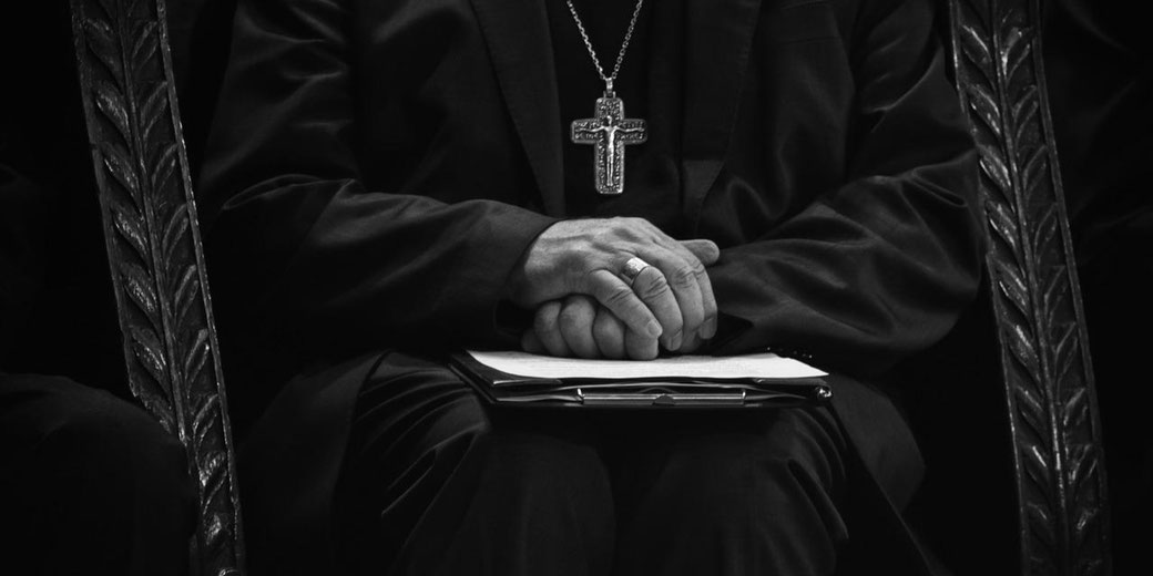 A person in dark clergy attire sits with hands clasped over a folder, wearing a large cross necklace and a ring, framed by a chair with a leaf-patterned design.