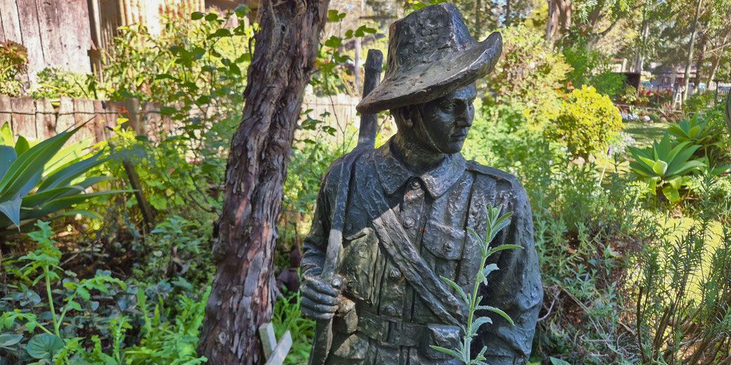 A soldier statue wearing a slouch hat and holding a rifle is placed in a garden surrounded by dense greenery.