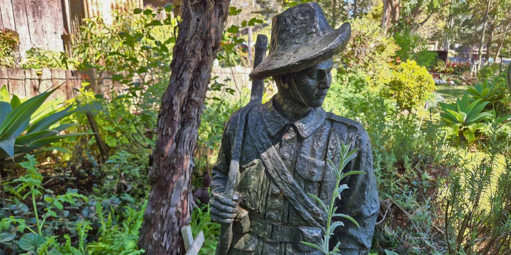 A soldier statue wearing a slouch hat and holding a rifle is placed in a garden surrounded by dense greenery.