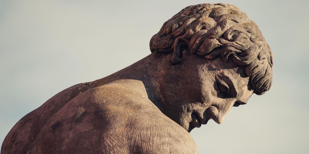 A weathered statue of an ancient Greek man with a muscular build and curly hair. His head is tilted downward, conveying introspection.