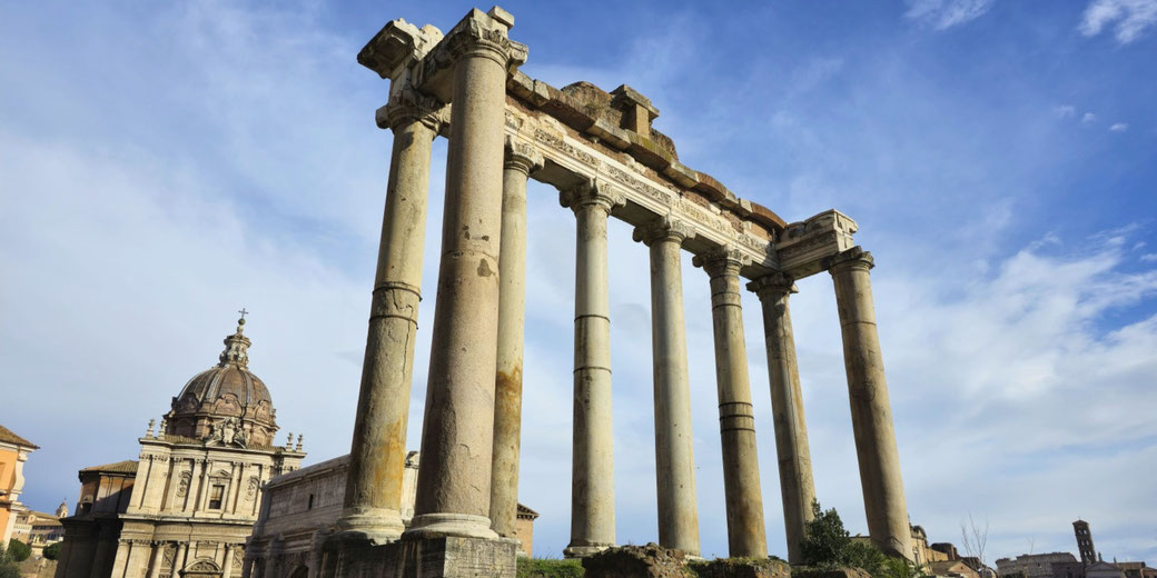 The ruins of the Temple of Saturn in the Roman Forum, Rome. The remaining tall columns and entablature stand against a blue sky, with historic buildings visible in the background.