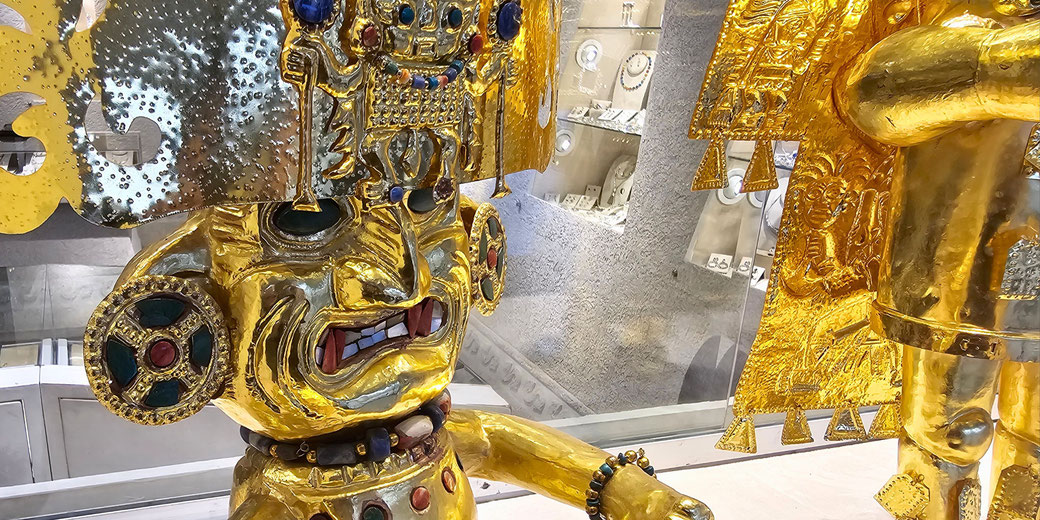 Golden pre-Columbian warrior figure with elaborate headgear, gemstone eyes, and intricate chest and ear decorations displayed in a museum setting.