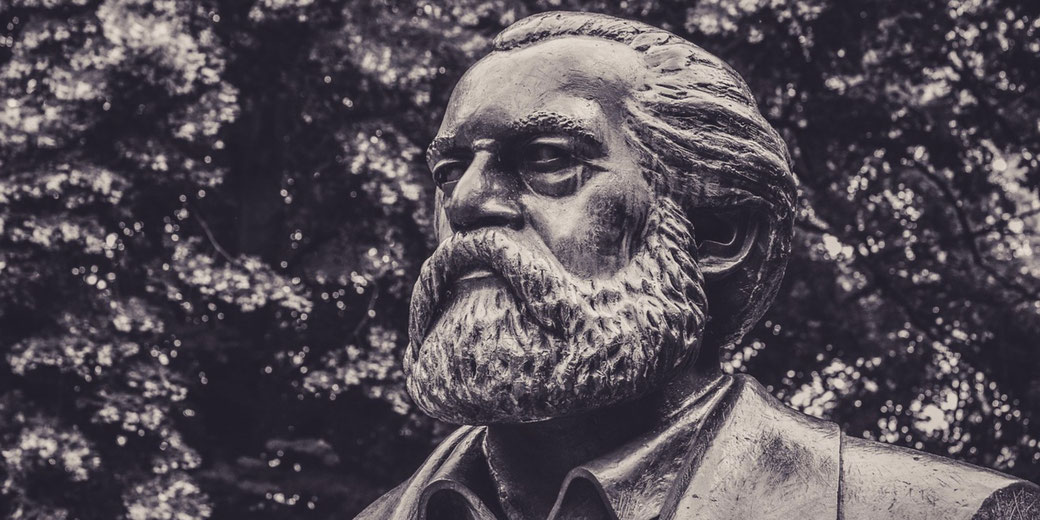 Close-up of a bronze statue of a bearded man with swept-back hair, set against a background of trees.