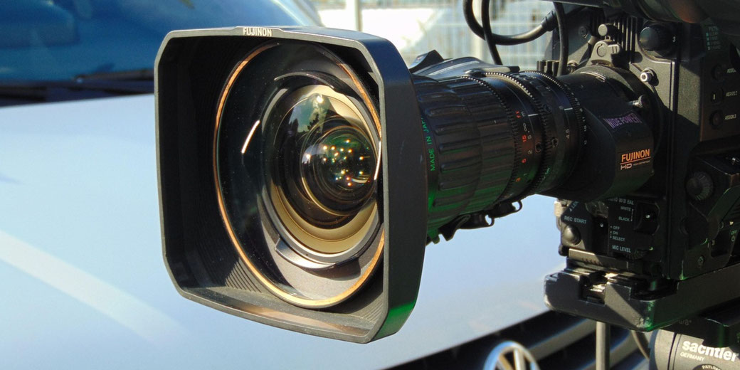 Close-up of a Fujinon HD video camera lens on a tripod, capturing outdoor reflections.