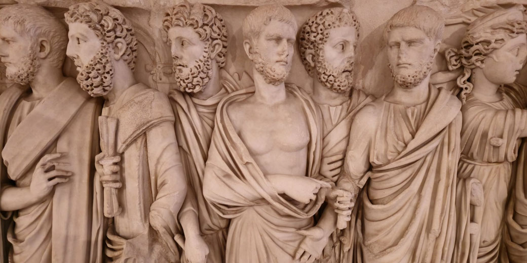 Relief sculpture of seven robed male figures with bearded faces, some holding scrolls, standing closely in a row with detailed expressions and garments.