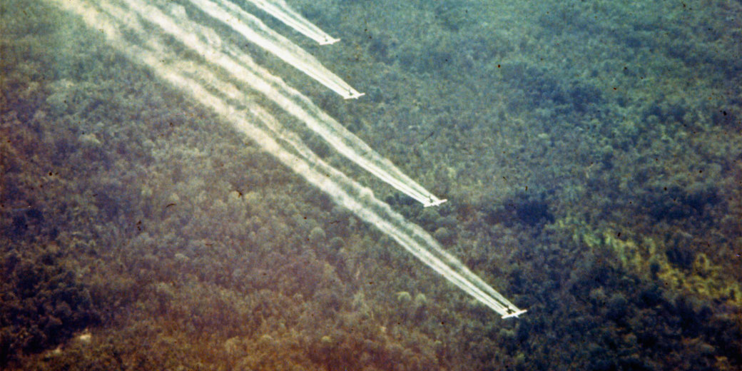 Four planes flying in formation, releasing trails of herbicide over a dense forest, likely depicting the use of Agent Orange during the Vietnam War.
