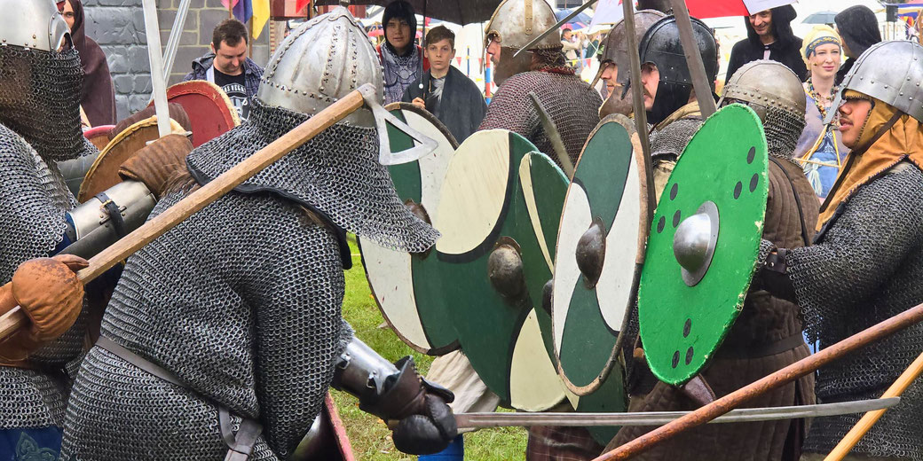 Group of reenactors in chainmail and helmets clashing with swords and shields during a staged medieval battle, with onlookers watching.