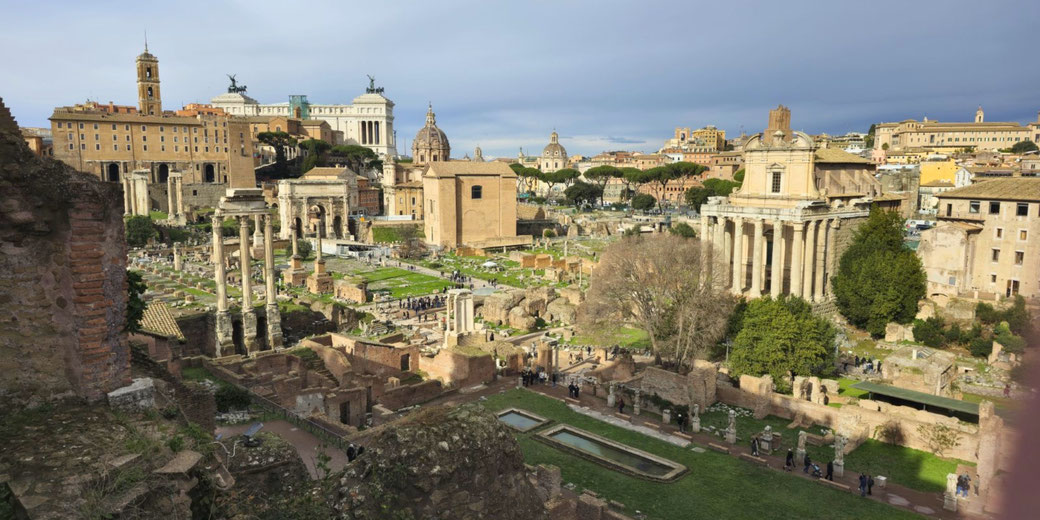 Panoramic view of the Roman Forum with ancient ruins, columns, and temples surrounded by modern city buildings.