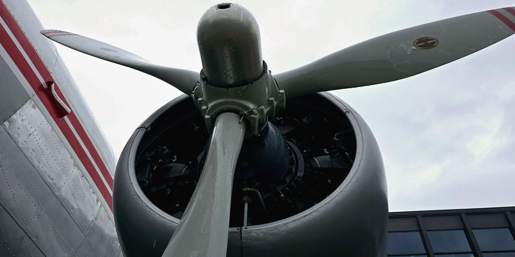 A close-up of a vintage airplane's propeller and radial engine. The sleek metallic blades are attached to the engine, with a red-and-white-striped fuselage visible on the left under a cloudy sky.