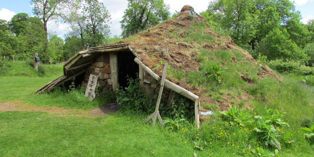A grass-roofed ancient-style earth hut with wooden supports sits nestled in a green, wooded area.