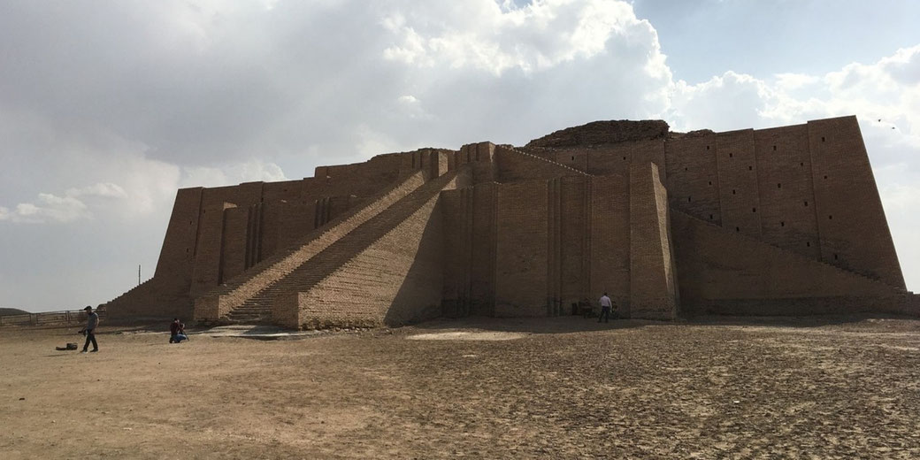 Massive ancient brick ziggurat with steep staircases stands under a cloudy sky, surrounded by dry, flat ground and a few visitors.