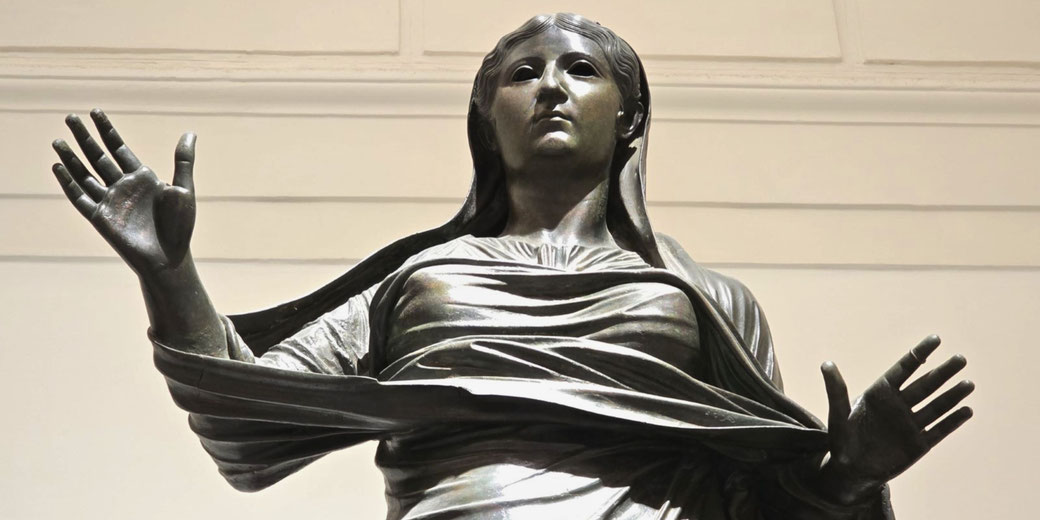 Bronze statue of a woman with raised hands, draped in flowing robes, gazing solemnly forward in a gesture of reverence or address.
