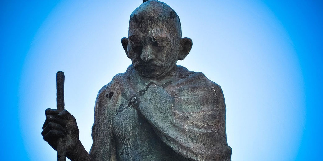 Statue of Mahatma Gandhi holding a walking stick, head bowed, with a rough surface and calm posture set against a clear sky, conveying resolve, humility, and leadership.