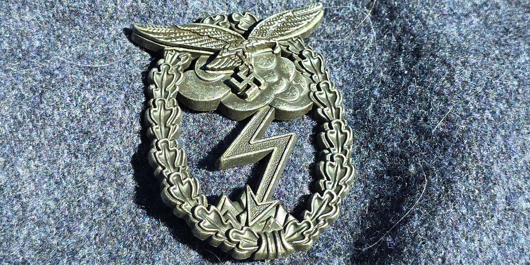 Metal badge featuring an eagle clutching a swastika above a lightning bolt, surrounded by an oak leaf wreath, displayed on a woolen fabric background.