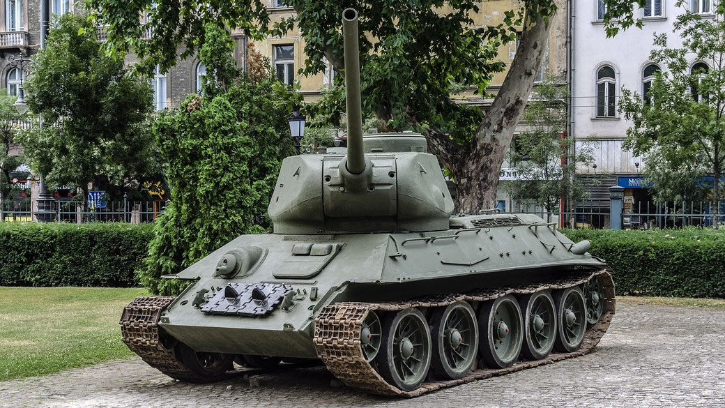 A green WWII-era tank is displayed on a cobblestone area in a park with buildings and trees in the background.