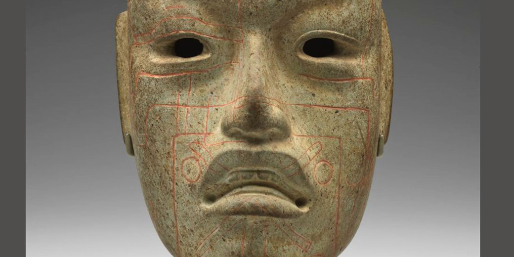 A carved stone mask with hollow eyes and a solemn expression, featuring intricate red geometric and symbolic engravings. Likely of Mesoamerican origin, it has a smooth, weathered surface with a ritualistic design.