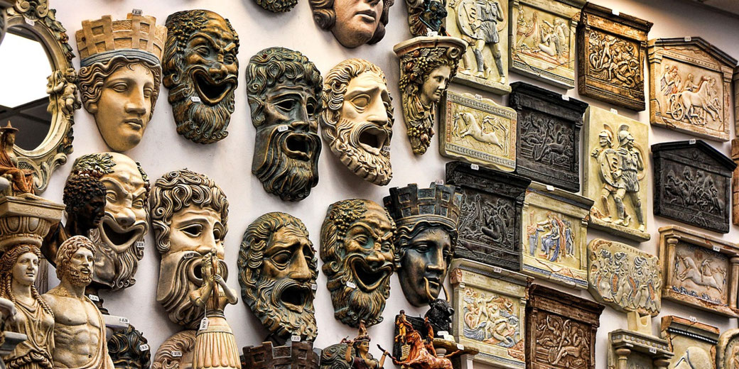 Display wall filled with colorful theatrical masks, relief plaques, and small classical sculptures, representing ancient Greek and Roman art themes.